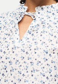 Κοντινή λήψη λευκού μπλουζιού με μικρό μπλε λουλουδάτο σχέδιο, με λαιμόκοψη με βολάν που δένει με φιόγκο και άνοιγμα σε σχήμα κλειδαρότρυπας.