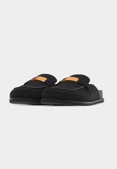 Ζευγάρι μαύρα loafers χωρίς κορδόνια με χαμηλά τακούνια και μεταλλικές λεπτομέρειες σε χρυσό τόνο στο πάνω μέρος, εμφανιζόμενα πάνω σε απλό ανοιχτόφωτο φόντο.