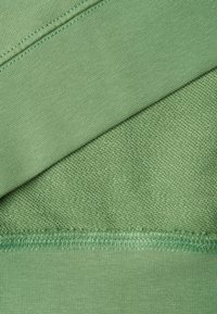 Gros plan sur un tissu vert clair montrant une surface tricotée texturée avec des coutures et des détails de piqûres visibles.