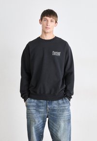 Schwarzer Sweatshirt mit Rundhalsausschnitt und gerippten Bündchen, mit weißem Text "Fantasy people" auf der Brust. Getragen mit blauen Jeans.