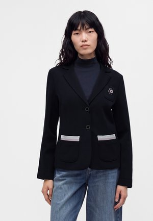 Kvinde med langt mørkt hår iført en sort blazer med to forlommer, et blomstret mærke, en mørk rullekrave og blå jeans.