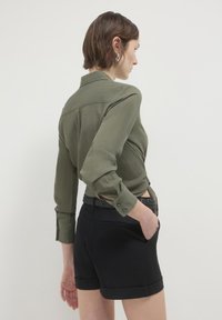 Camicia verde oliva con bottoni in tessuto texture, maniche lunghe, polsini arrotolati e una vita nera a contrasto abbinata a pantaloni corti neri sartoriali.