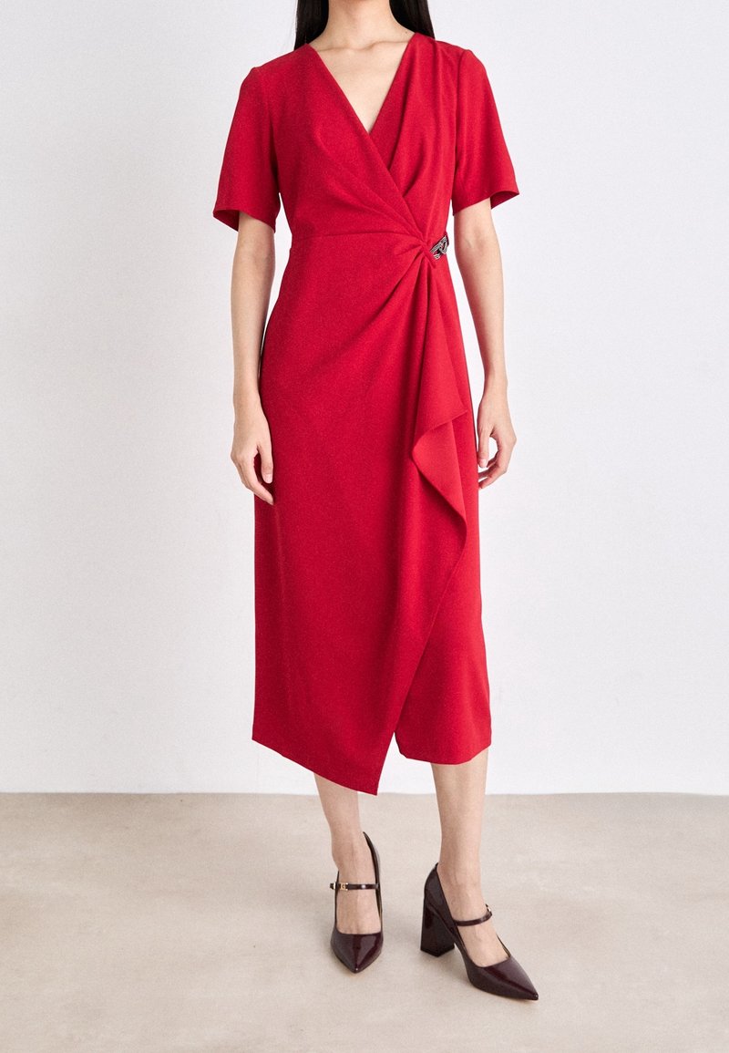 Femme portant une robe midi portefeuille rouge avec manches courtes et détail de volants, associée à des talons hauts sombres à bout pointu avec des brides à la cheville.