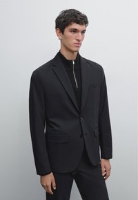 Giacca nera aderente con collo a risvolto, chiusura con un solo bottone e due tasche frontali. Abbinata a un top in maglia nera con zip sottostante.