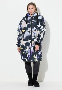 Flerfärgad puffjacka med ett stjärnmönster, svart bas, dragkedja fram, huva och elastiska ärmslut; kombinerad med mörka jeans och svarta stövlar.