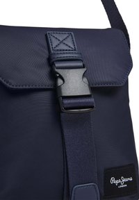 Borsa in nylon blu navy con chiusura magnetica, tracolla regolabile e tessuto testurizzato. Presenta un'etichetta con logo e cuciture rinforzate.