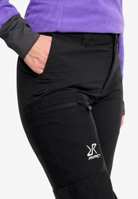 Person som bär svarta byxor med en ficka med dragkedja på låret och en lila fleeceströja, hand delvis i framsidan av fickan.