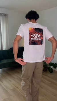 Person, die den Saum eines weißen Umbro-T-Shirts mit einer orange-schwarzen Grafik auf der Rückseite in einem Wohnzimmer anpasst.