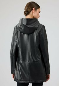 Giubbotto in pelle nera con cappuccio, taglio dritto e tasca laterale con zip. Texture liscia con dettagli delle cuciture sul retro.