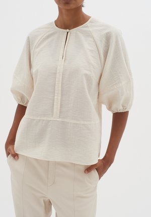 Vrouw draagt een crèmekleurige blouse met half opgezette mouwen en een keyhole-halslijn, gecombineerd met lichtbeige broek, handen in de zakken.