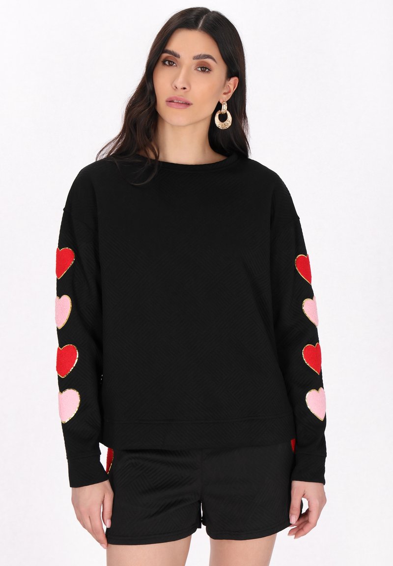 Zwarte pullover trui met lange mouwen, voorzien van geborduurde rode en roze hart applicaties op beide mouwen en een textuur patroon.