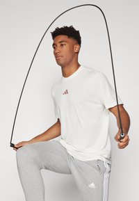 Hvid atletisk T-shirt med rødt logo; grå joggere med hvide striber; person der holder et sort sjippetov i en klar stilling.