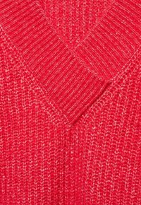 Pull en tricot rouge avec un design en V, présentant un motif texturé et des accents côtelés. Le fil a un effet subtil bicolore.