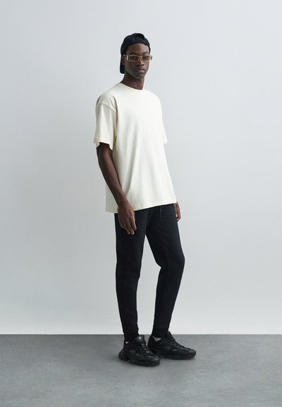 Beige loose-fitting t-shirt, black slim trousers, black sneakers with textured detail, and a black cap. Clear background.