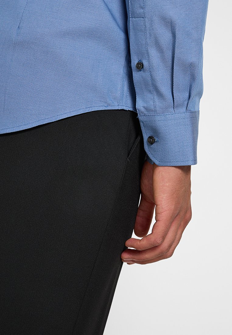 Chemise texturée bleu clair à manches longues, avec des poignets boutonnés, et une jambe de pantalon noire ajustée visible. Tissu lisse avec un motif subtil.