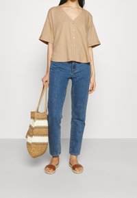 Blusa beige de manga corta con escote en V y botones en la parte delantera, combinada con jeans azul de pierna recta y sandalias marrones, sosteniendo un bolso de mimbre.