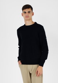 Maglione nero lavorato a maglia con scollatura rotonda, polsini e orlo a coste, con un dettaglio del logo sulla manica sinistra. Tessuto liscio.