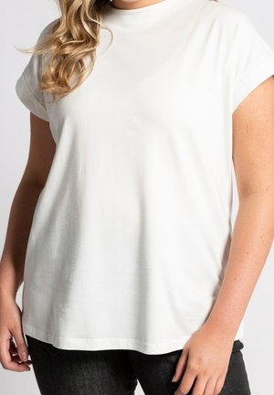 Kobieta w gładkim, białym, krótko rękawowym T-shircie i czarnych spodniach, z częściowo widocznymi blond kręconymi włosami na ramieniu.