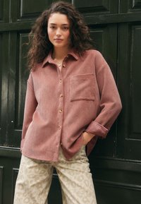 Camicia rosa texturizzata con bottoni e taschino sul petto, indossata sopra pantaloni chiari con stampa leopardata. La camicia presenta bottoni in legno e polsini arrotolati.