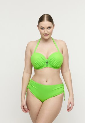 Vrouw met naar achteren getrokken haar die een neon groene halterbikini draagt met gerimpelde, hoge taillebroekjes, staand tegen een effen witte achtergrond.