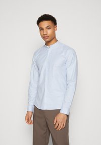 Camicia azzurro chiaro a maniche lunghe con colletto a banda e chiusura frontale con bottoni. Il tessuto sembra liscio con una texture sottile. Visibili pantaloni marroni.