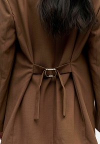 Bruine op maat gemaakte jas met een gestructureerde stof, een riem met een metalen gesp aan de achterkant en een gestructureerd ontwerp met plooien.