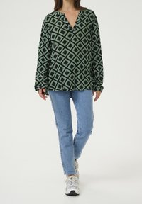 Camisa de manga larga negra y verde con un patrón de diamantes, con escote en pico y corte relajado, combinada con jeans de mezclilla azul claro y zapatillas.