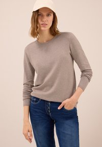 Žena v béžovém svetru s dlouhým rukávem, modrých džínech s knoflíky a bílém kšiltovce, stojící s jednou rukou v kapse na béžovém pozadí.