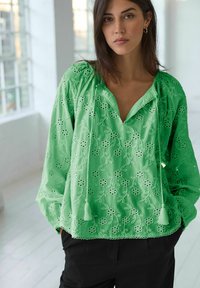 Blusa verde ricamata con motivi floreali, scollatura a V e dettagli con frange. Tessuto leggero con maniche lunghe e una vestibilità comoda.
