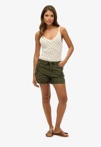 Débardeur en tricot blanc avec un motif en losanges associé à un short cargo vert olive. Le mannequin porte des sandales plates marron, les mains dans les poches du short.