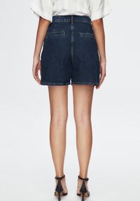 Tmavě modré džínové šortky s vysokým pasem, dvěma zadními kapsami a ohrnutým lemem, doplněné černými sandály na vysokém podpatku, zobrazené zezadu.