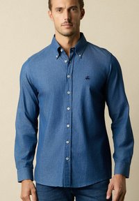 Denim overhemdbloes in diepblauw, met een gestructureerde kraag, lange mouwen en een subtiele geborduurde logo op de borst.