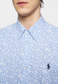 Jasnoniebieska koszula na guziki z kwiatowym wzorem wykonana z tkaniny o fakturowanym wzorze. Posiada klasyczny kołnierzyk oraz haftowane logo na piersi.