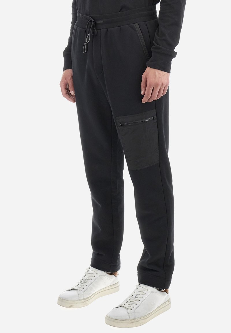 Zwarte joggingsbroek van een gladde, rekbare stof. Voorzien van een ritszak aan de zijkant en een elastische tailleband met trekkoord. Gepaard met witte sneakers.