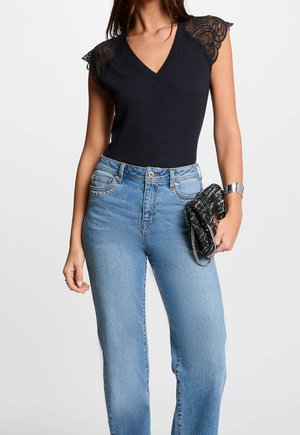 Mujer con top negro sin mangas con hombros de encaje y pantalones vaqueros azules de cintura alta, sosteniendo un bolso clutch negro texturizado.