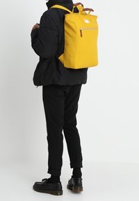 Zaino giallo con forma rettangolare, tasca con zip e dettagli in pelle. Indossato da una persona con un giubbotto imbottito nero e pantaloni.