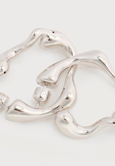 Ασημένια σκουλαρίκια χειροποίητου σχήματος με κυματιστό, οργανικό σχέδιο και κλείσιμο με καρφίδα, σε λευκό φόντο.