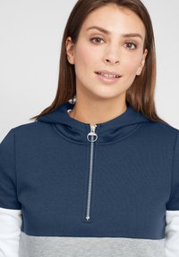 Felpa con zip di colore blu navy, grigio e bianco. Presenta un cappuccio, maniche corte e una cerniera metallica. Realizzata in morbido tessuto testurizzato.