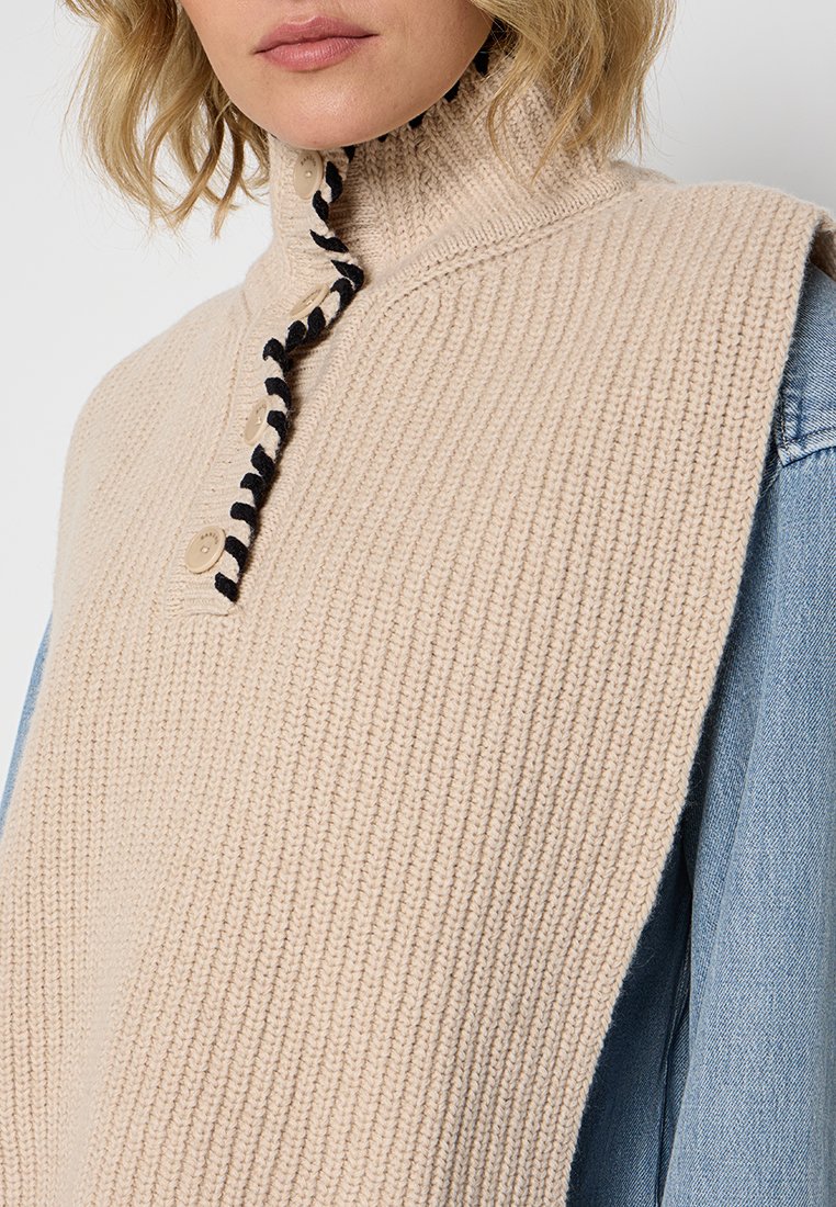 Beige gestrickte Weste mit hohem Kragen, ausgestattet mit einem Knopfverschluss und einem kontrastierenden schwarz-weißen geflochtenen Krawattendetail.