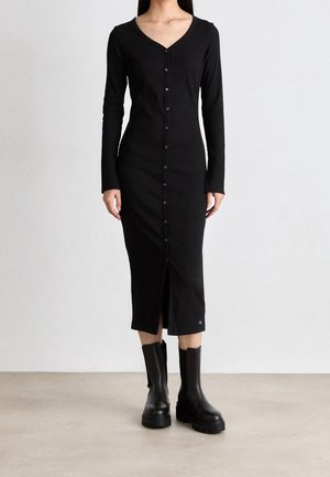 Kobieta ubrana w dopasowaną czarną midi sukienkę na guziki z długim rękawem i dekoltem w serek oraz czarne botki, stojąca na jednolitym tle.