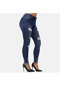 Vaqueros ajustados en denim azul oscuro con rasgaduras desgastadas; presentan dobladillos deshilachados y costuras decorativas a lo largo de los lados, combinados con tacones negros.