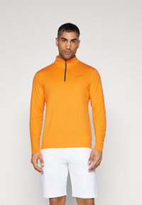 Oranžni dolg rokav z zadrgo, izdelan iz gladkega in lahkega materiala. Ima visok ovratnik in črn logo Calvin Klein na prsih.