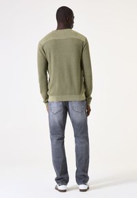 Mann in grünem strukturiertem Pullover, verblassten blauen Jeans und weißen Sneakern, der mit dem Rücken zur Kamera vor einem weißen Hintergrund steht.