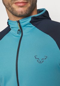 Tyrkysová bunda na zips s námorníckymi ramennými panelmi. Obsahuje vysoký golier, zlatý zips a nenápadné logo pri leme. Mäkká, štruktúrovaná tkanina.