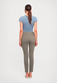 Kvinne med brun hestehale iført en blå kortermet topp, olivengrønne smale jeans og beige hæler står med ryggen til foran en ensfarget hvit bakgrunn.
