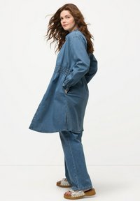 Abito in denim con maniche lunghe, vita arricciata e tasche laterali. Colore azzurro chiaro, vestibilità casual e lunghezza media. Indossato con pantaloni in denim e sandali.