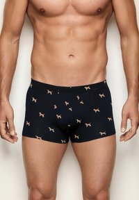 Mann in dunkelblauen Boxershorts mit kleinen Beagle-Hundemustern, der vor einem schlichten hellen Hintergrund steht.