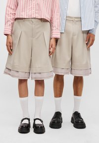 Beežid šortsid voldikutega, kihiline äär, kus on läbipaistev helesinine alumine osa. Kandmisest erineva värvi jalatsitega ja valgete sokkidega.