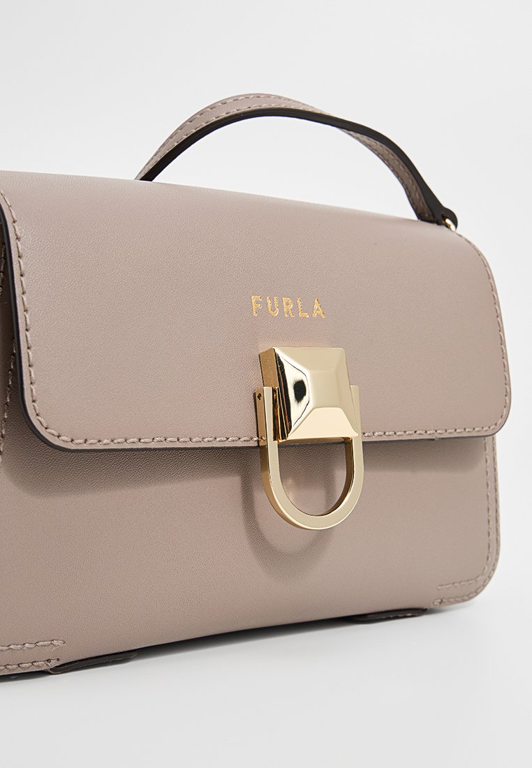 Beige Lederhandtasche mit einem oberen Griff, versehen mit einem glänzenden goldenen Metallverschluss und dem in Gold eingeprägten Logo "FURLA" auf der Vorderseite.
