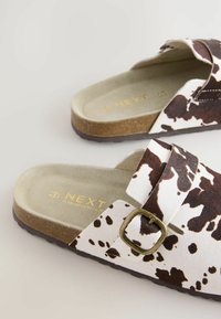 Sandalias de dedo con estampado de vaca en blanco y marrón texturizado. Cuentan con una correa ancha, hebilla ajustable y una plantilla contorneada para mayor comodidad.